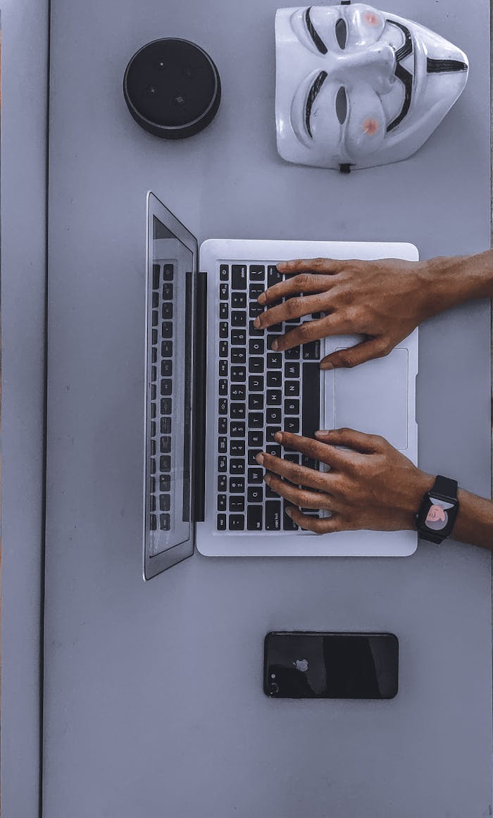 Top view of hands typing on a laptop beside a mask and smartphone.