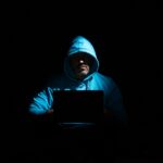 A person in a hoodie using a laptop in a dark setting, conveying mystery and technology themes.